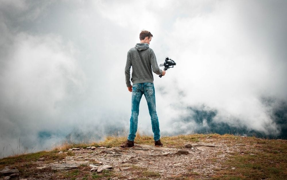 一个穿着牛仔裤的男孩站在一块岩石上，手里拿着一台专业相机，在拍摄他面前的云。