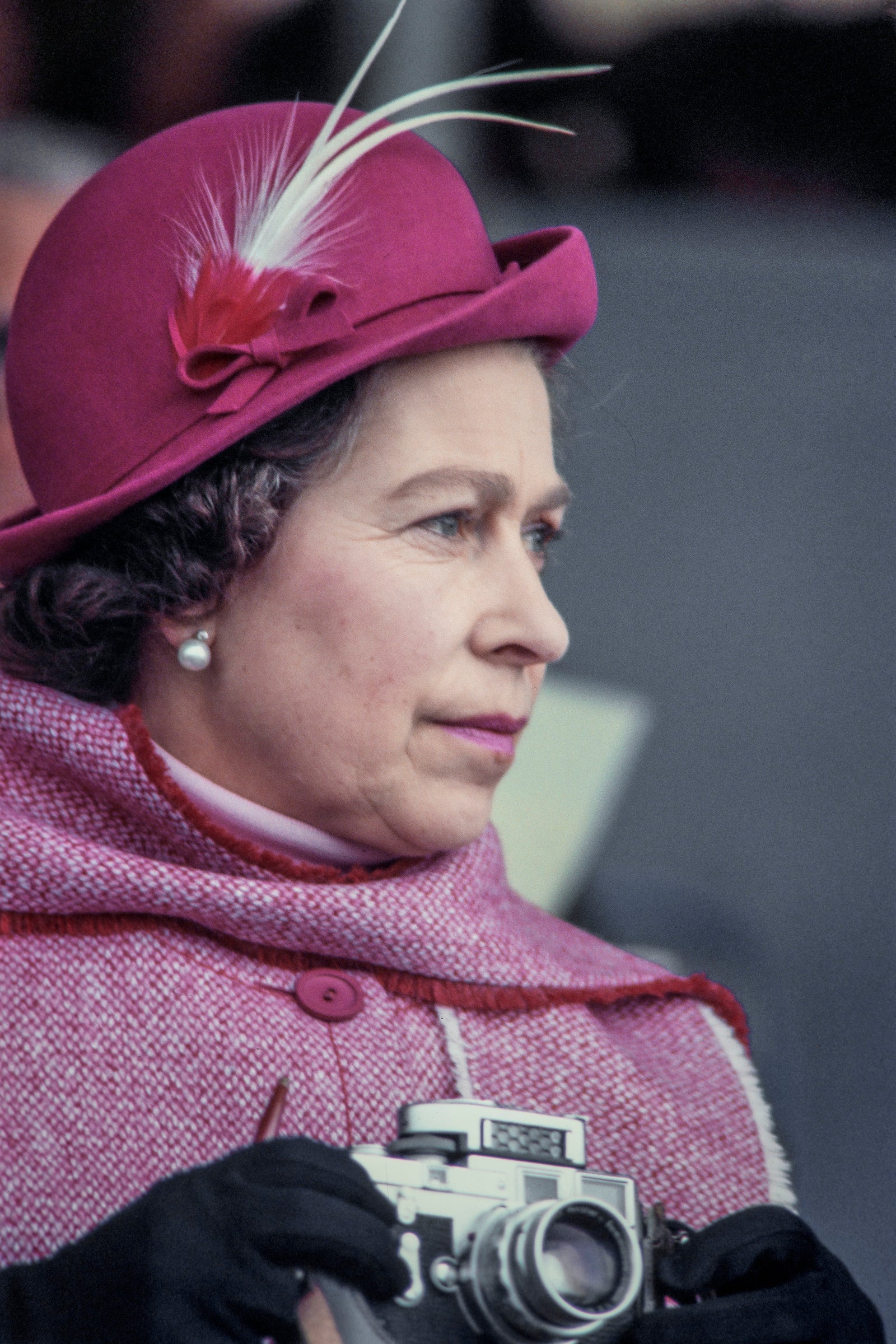 英国女王伊丽莎白二世的特写，她戴着淡红色的羽毛毡帽，披着羊毛斗篷，手持徕卡M3相机参加马术活动，摄于1975年左右的英国温莎。